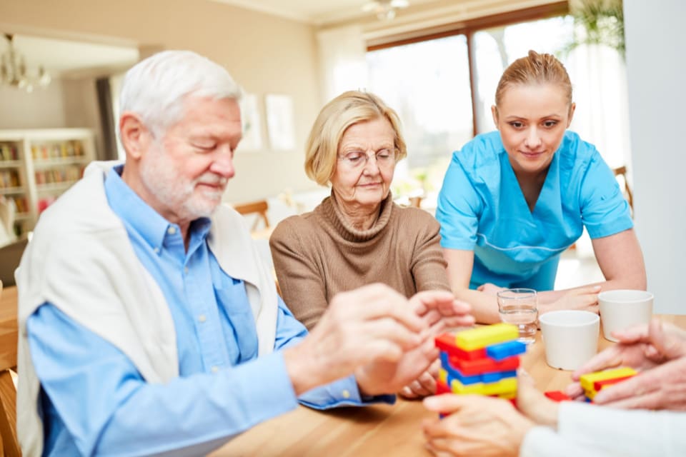 A caregiver watches a senior man and senior woman put together a building toy