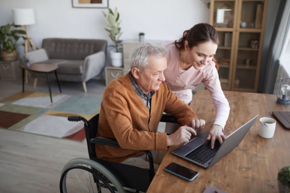 A caregiver helps a senior man in a wheelchair to use a laptop